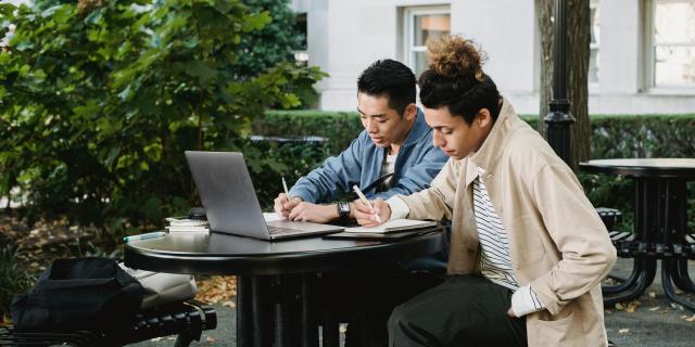 Two students sit at an outdoor table, taking notes, with a laptop open in front of them.